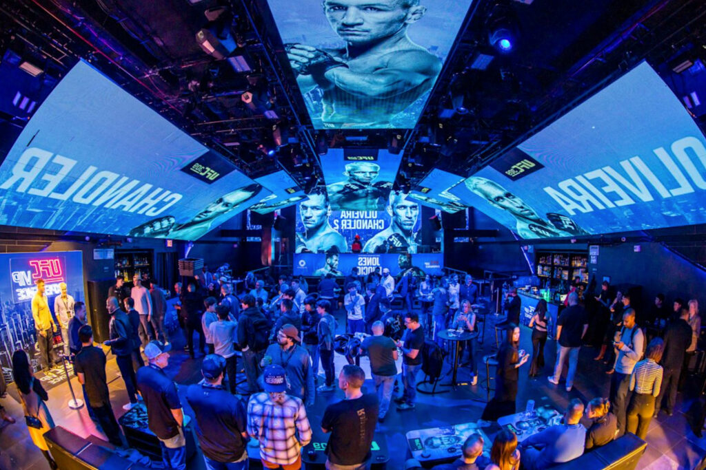 Guests mingle in a UFC VIP lounge lit blue beneath giant fighter-branded ceiling screens, part of the UFC VIP experience curated by Perfect.Live