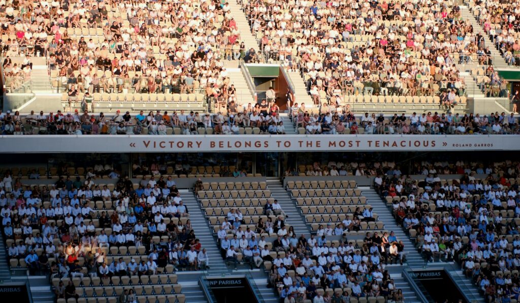 A packed daytime crowd in the stadium stands watching a professional tennis match