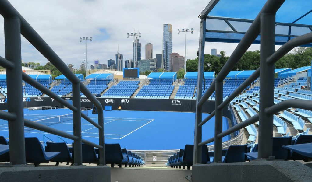 An empty blue hard tennis court seen from an elevated angle with a city skyline behind, daytime