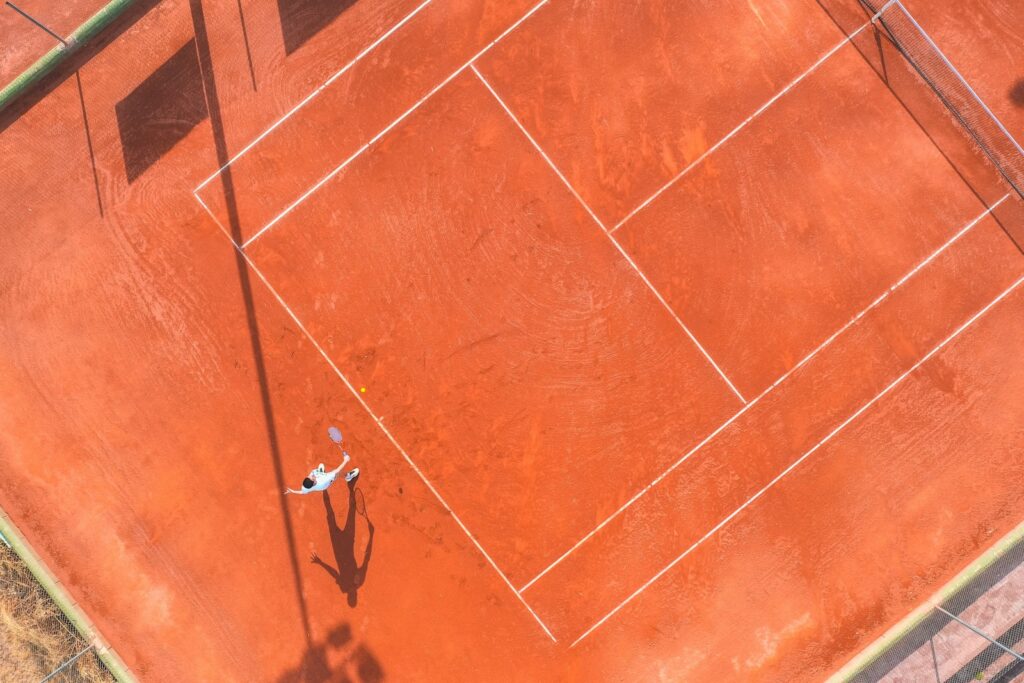 Aerial view of a red clay tennis court with a player and long shadow