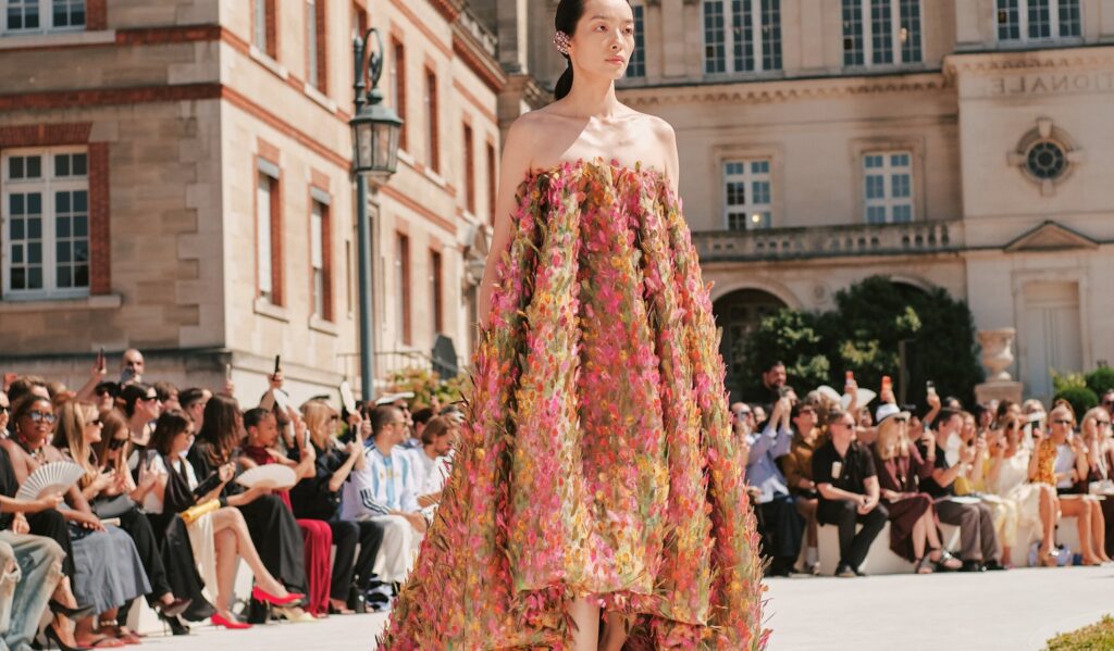 Paris Fashion Week 2026 — a model in a voluminous strapless gown covered in petal appliques on an outdoor Parisian runway, part of the fashion week access curated by Perfect.Live