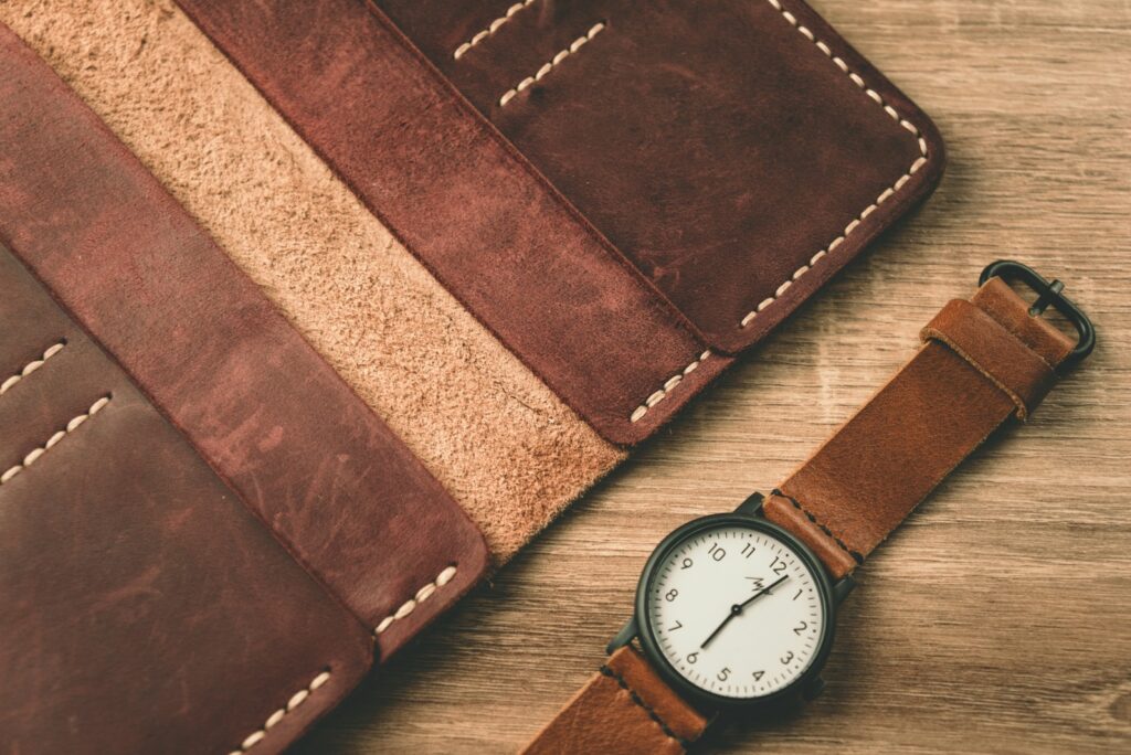 Hand-stitched leather wallet beside a leather-strap watch on a warm wooden surface