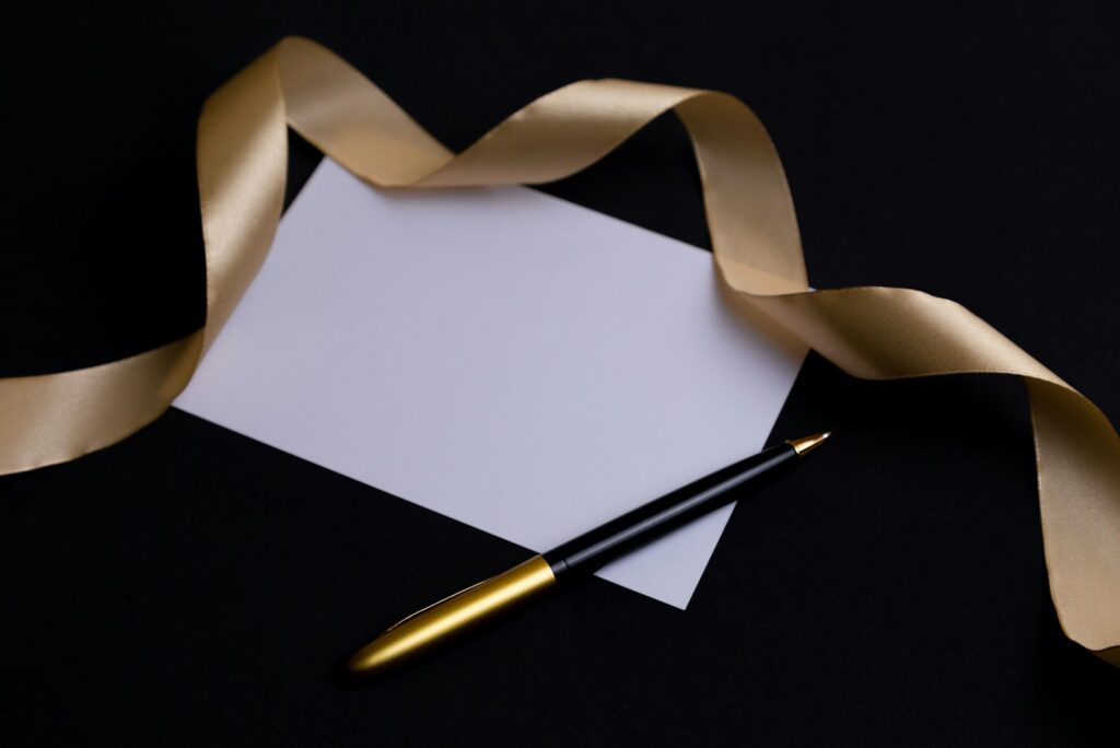 A blank white card, a gold satin ribbon, and a gold-and-black pen on a black surface.