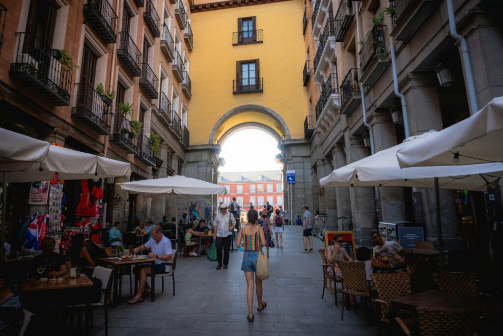 Café terraces beneath white awnings on a Madrid old-town street leading to a stone archway