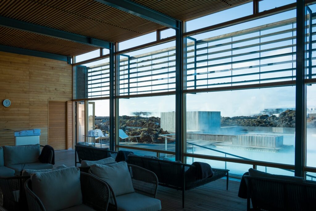 Quiet spa lounge with woven sofas facing tall windows and a steaming blue lagoon beyond