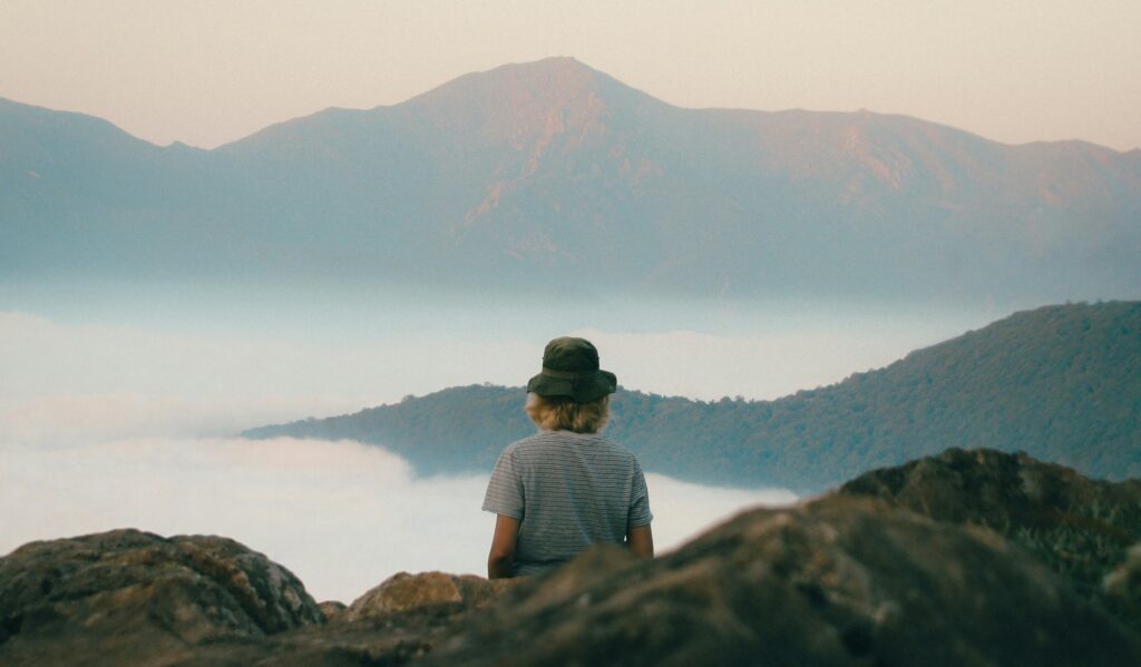 Meditation retreat: a guest seen from behind, seated in quiet reflection above a sea of cloud and mountains at dawn