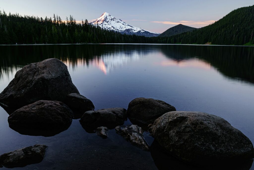 Still mountain lake at dusk with dark boulders in the foreground and a snow-capped peak beyond