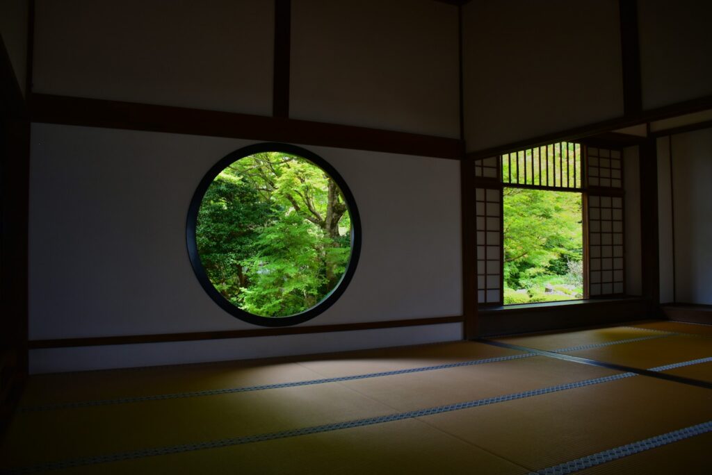 Empty tatami room with a round window opening onto green maple trees