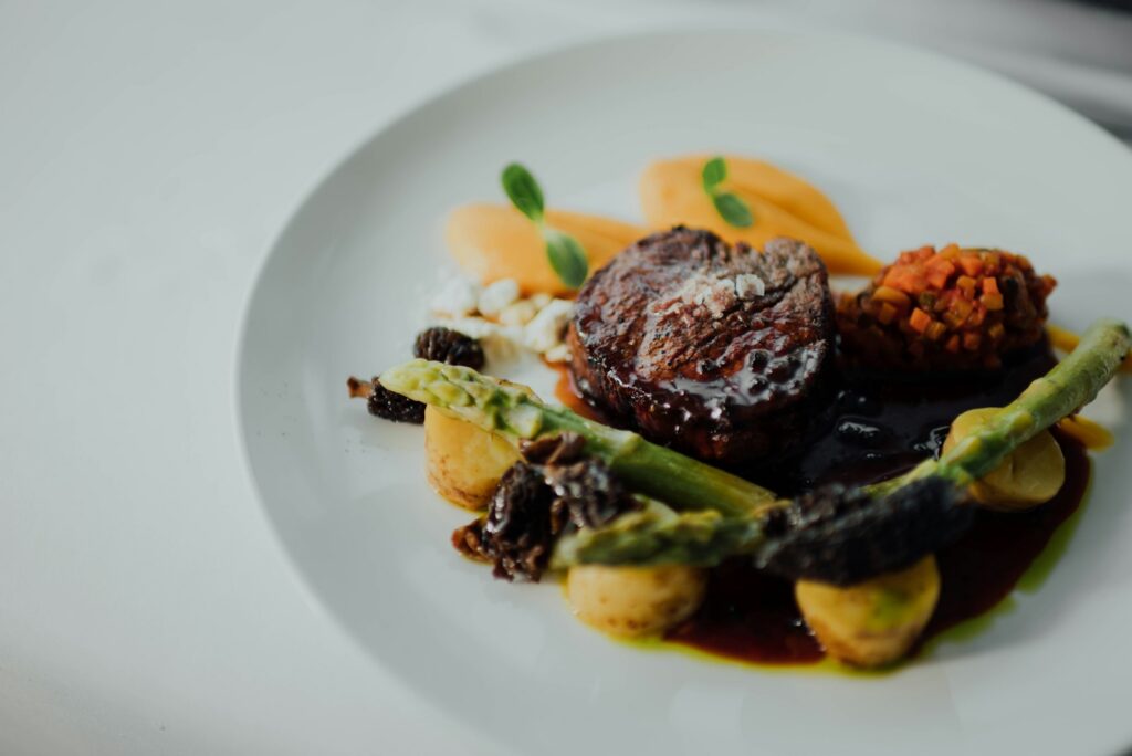 Close-up of a fine-dining course plated on white porcelain: seared beef fillet with morels, asparagus, baby potatoes, and carrot purée