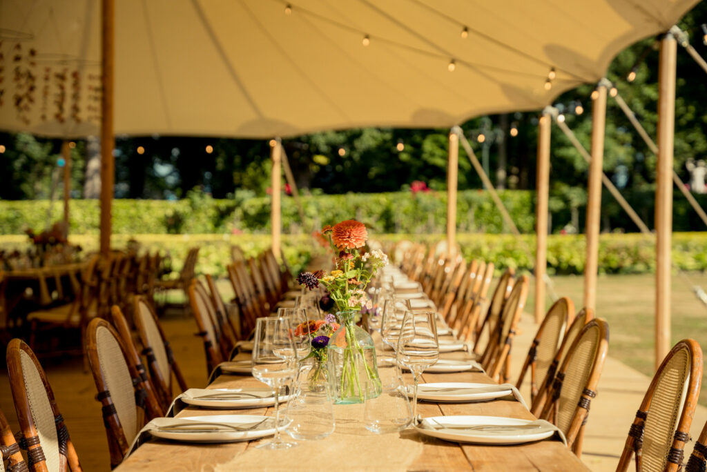 Long celebration table laid for guests under a marquee beside a garden hedge