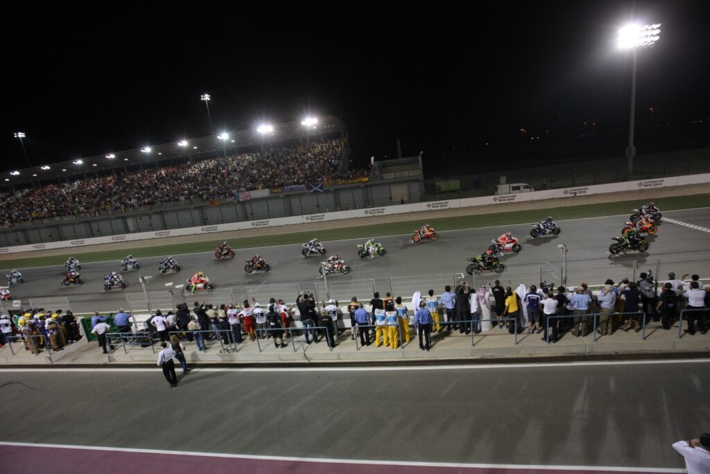 Riders launch from the grid at night under floodlights at Lusail circuit, with the main grandstand full for the Qatar Grand Prix
