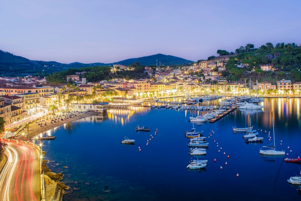 A Riviera harbour town lit up with yachts at anchor in the evening