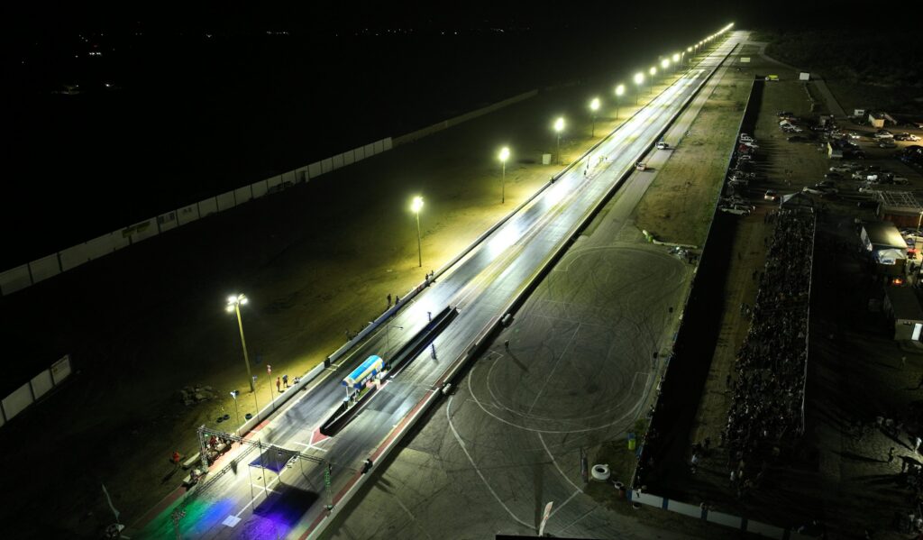 Aerial view of a floodlit race track at night, its long straight lit by tall lamps.