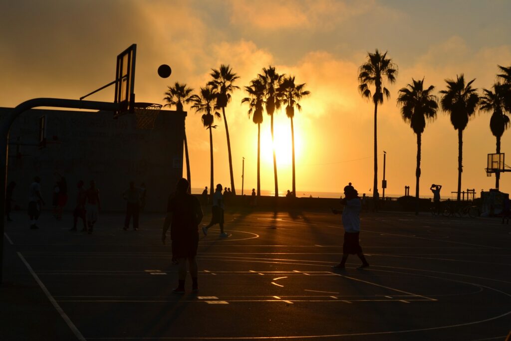 Silhouettes of people playing basketball on a beach court at sunset with palm trees in Los Angeles