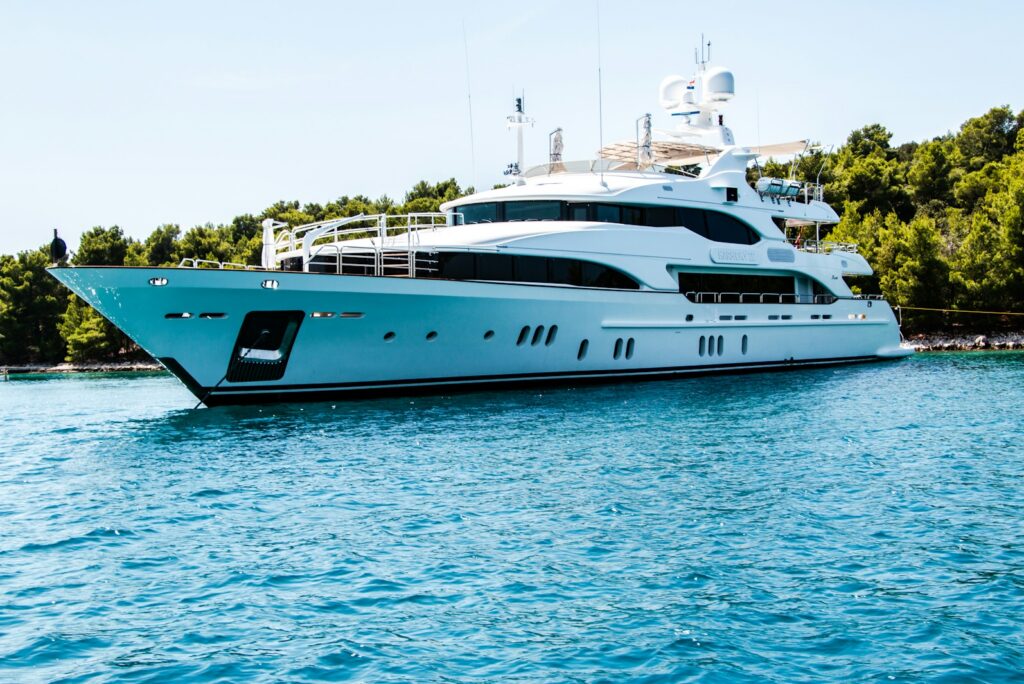 White motor yacht at anchor in calm water beside a green, tree-covered shoreline